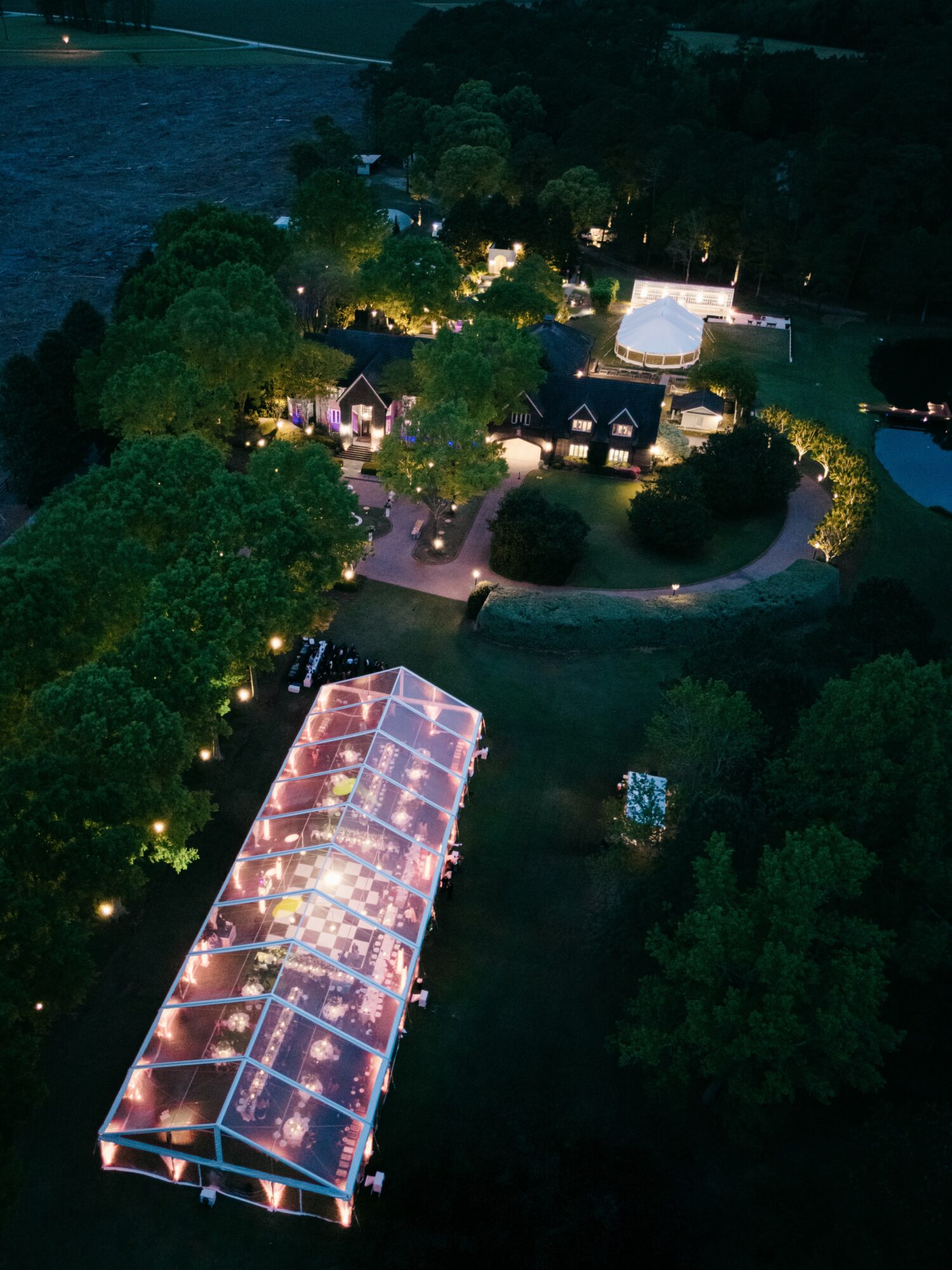 Night view of illuminated outdoor area with pathways, trees, and structures, including a large glass-roofed building, surrounded by greenery.