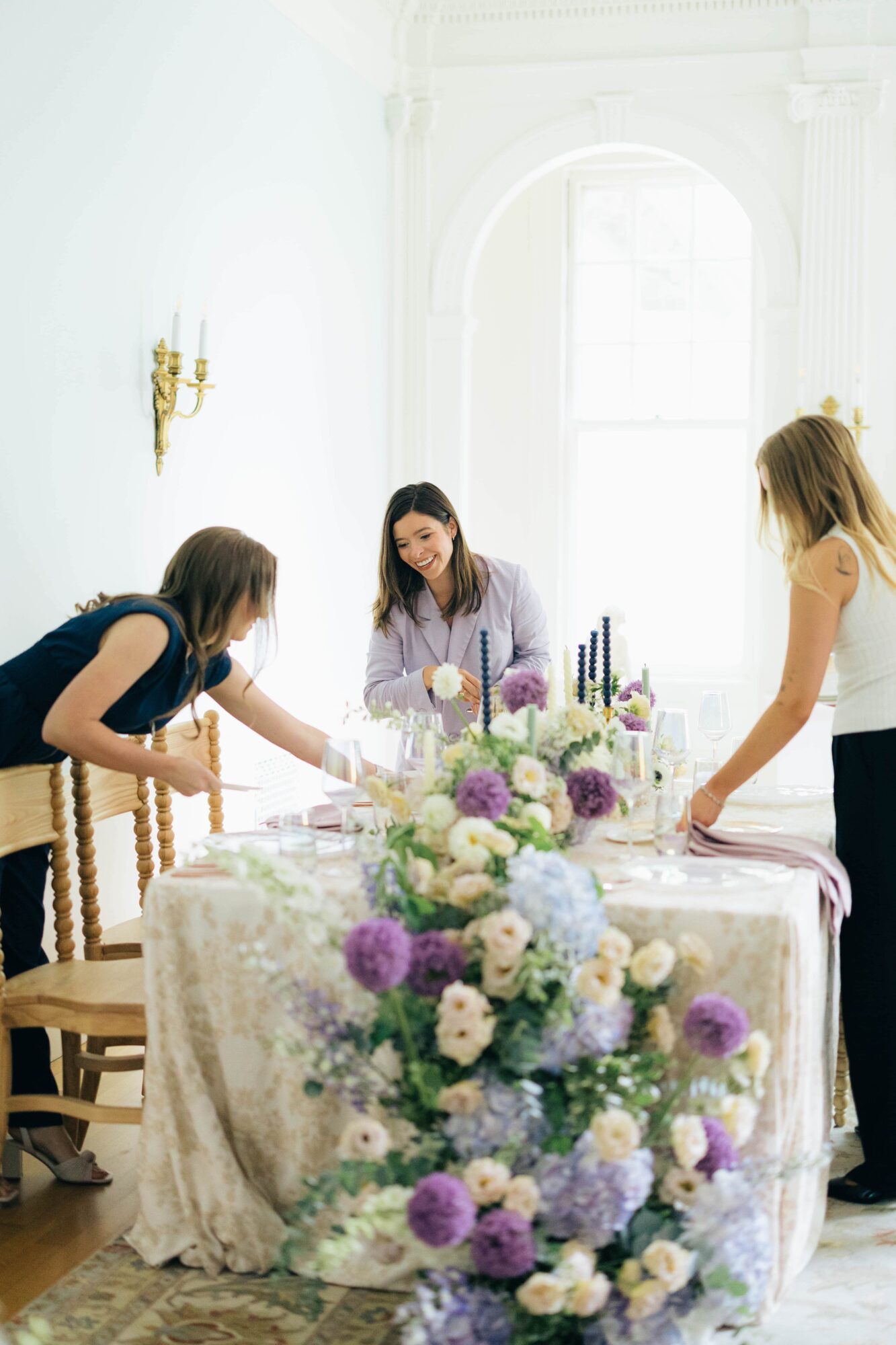 Three women prepare a table with a floral centerpiece in a bright room with large windows and white walls.