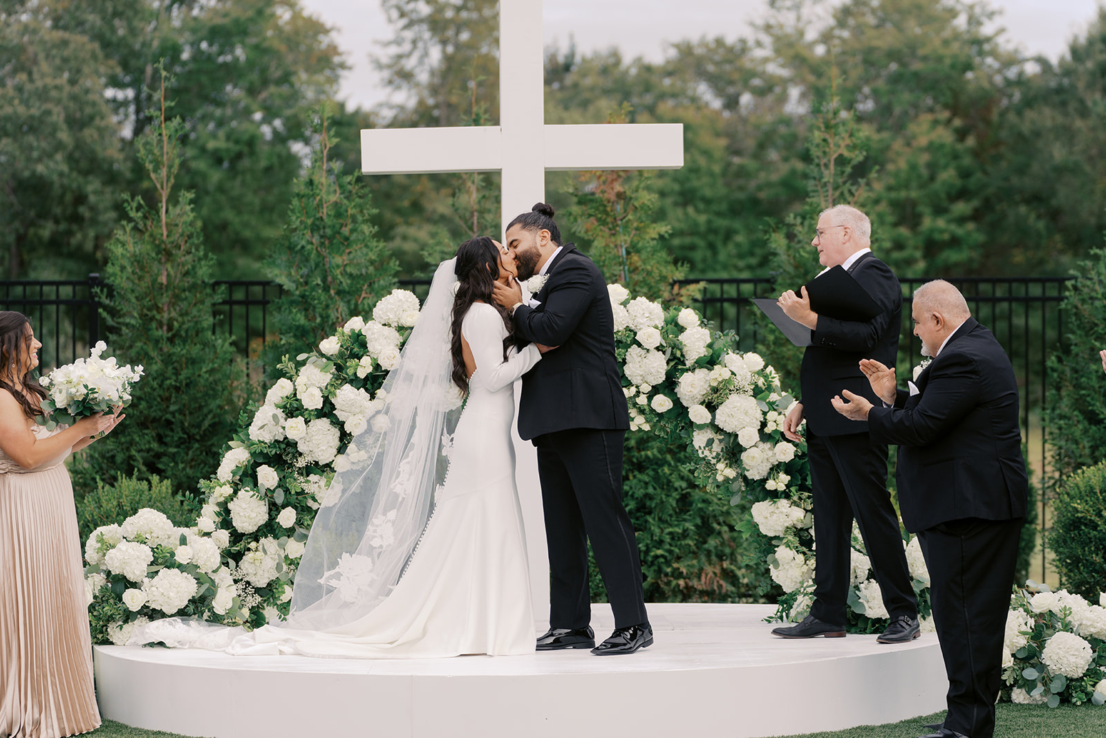 Couple kissing at wedding altar with large white cross and floral arrangements, surrounded by three people praying or holding flowers.