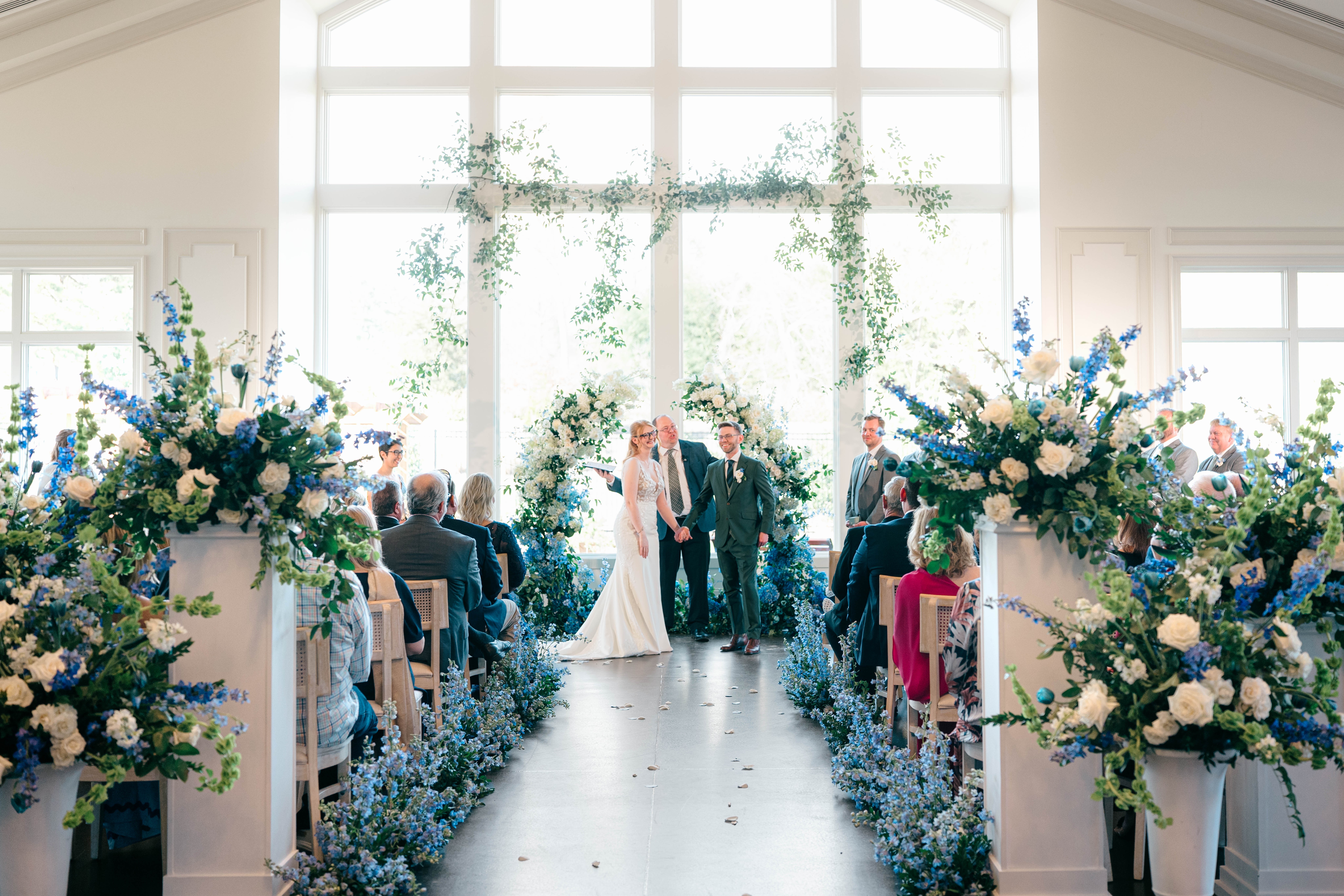 Wedding ceremony inside a bright, airy venue with large windows, floral arrangements, and guests seated facing the couple.