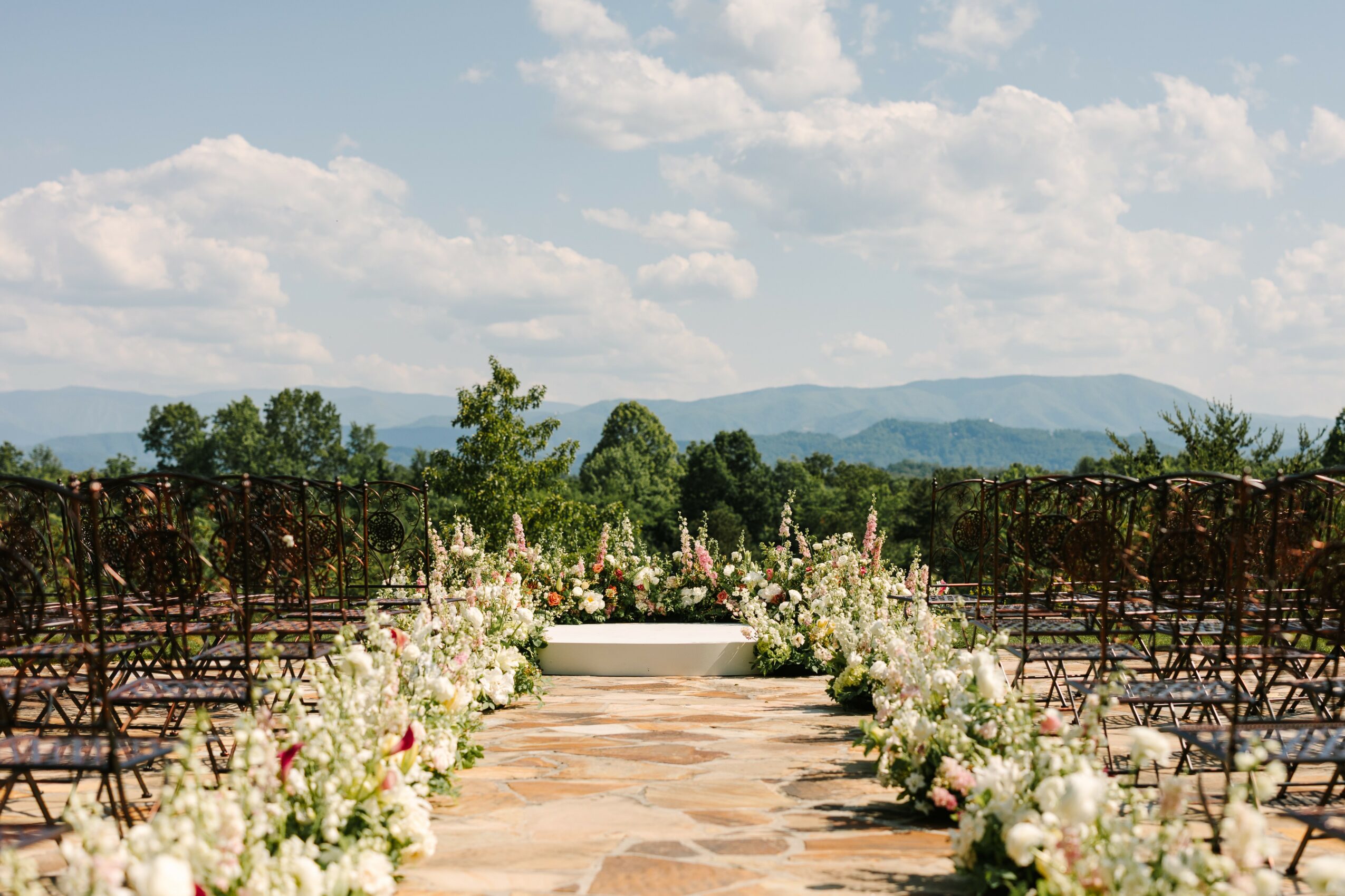 Outdoor wedding ceremony setup with rows of chairs and floral arrangements on a stone pathway, scenic mountains in background.