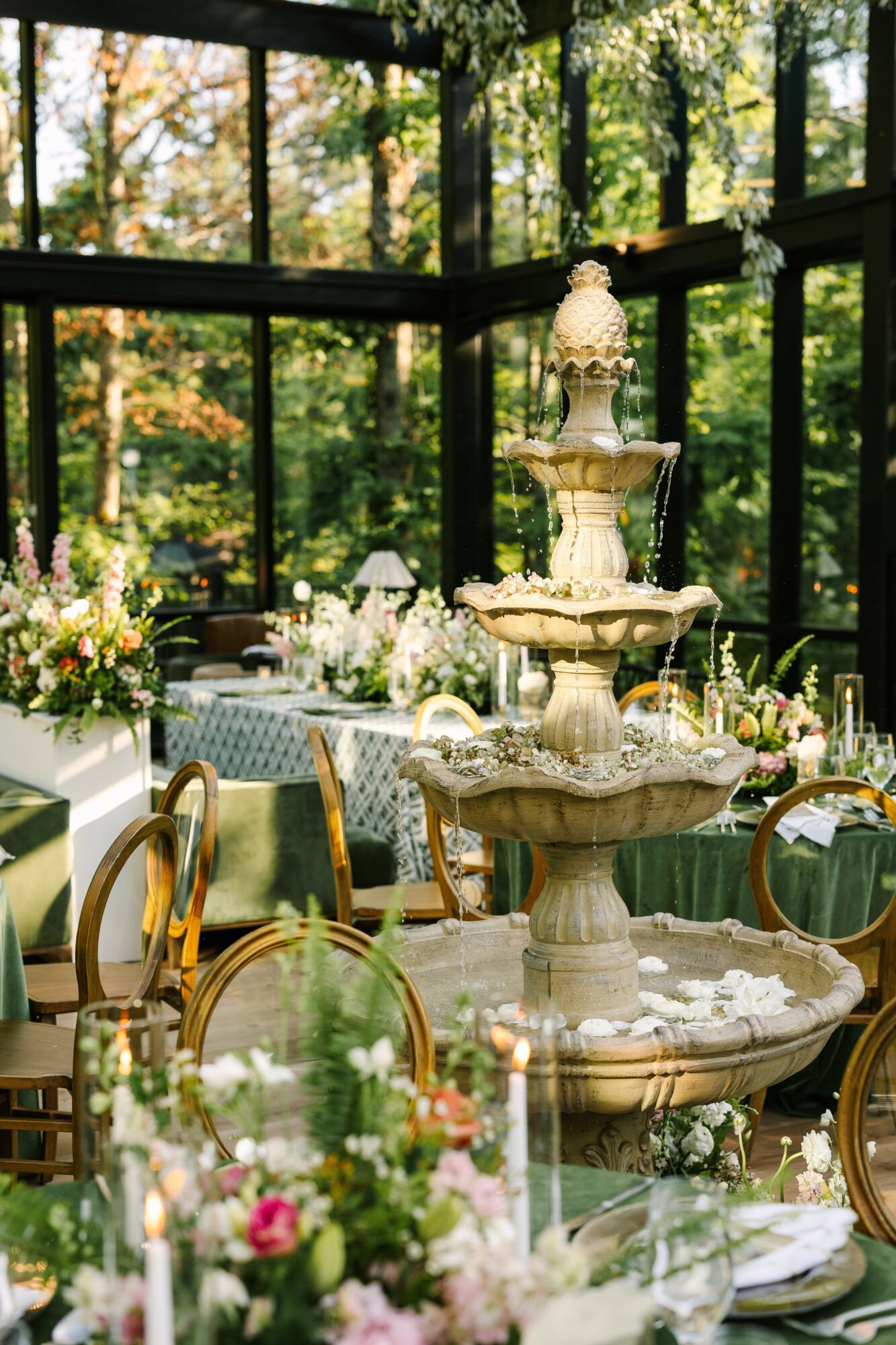 Elegant indoor space with a multi-tiered fountain, surrounded by floral arrangements and chairs, with large glass windows and greenery outside.