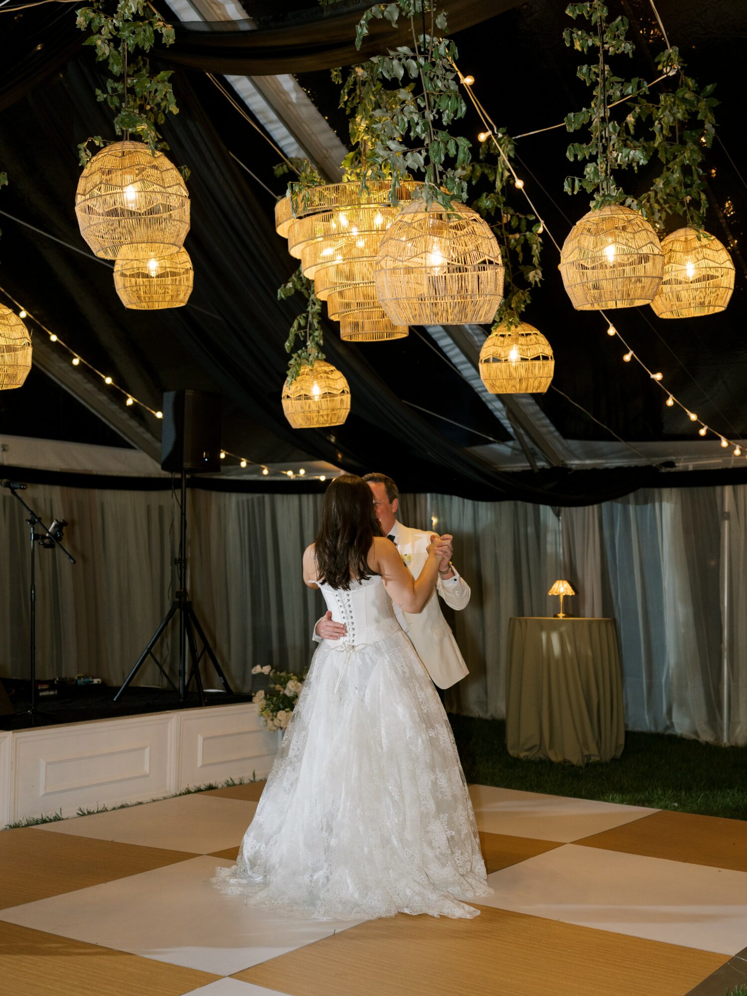 Couple dancing under hanging lanterns and string lights in an indoor venue with draped curtains and greenery.