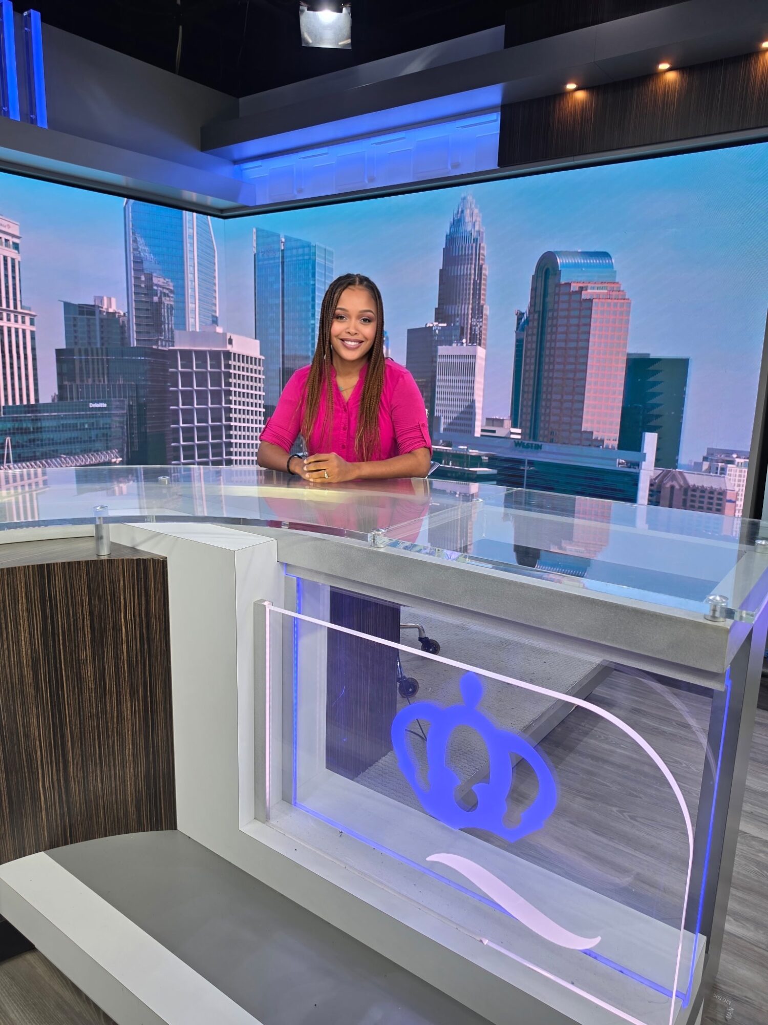 Female news anchor sitting at a news desk with a city skyline background, smiling, wearing a pink top.