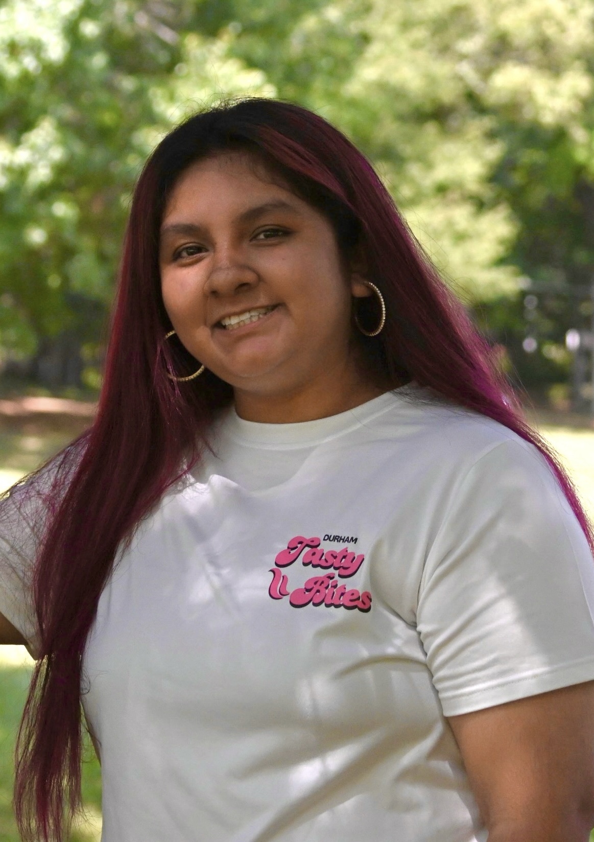 Smiling person with long dark hair and hoop earrings outdoors, wearing a white T-shirt with pink text, in a green park.