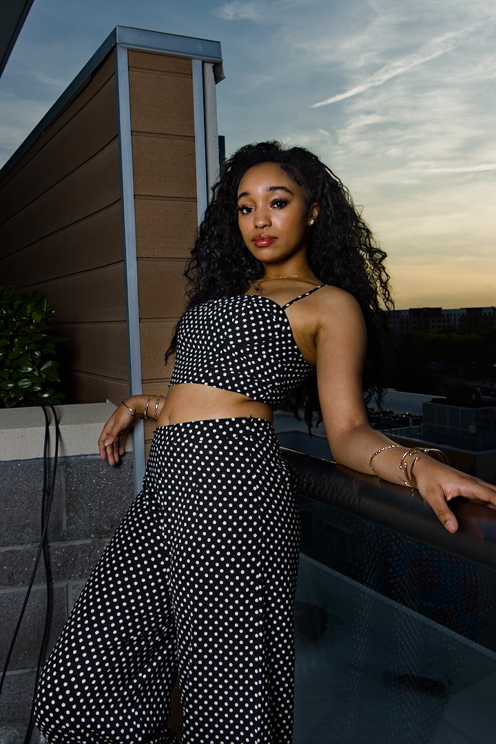 Young woman with curly hair in polka dot crop top and wide-leg pants, standing outdoors at sunset, leaning on a railing.