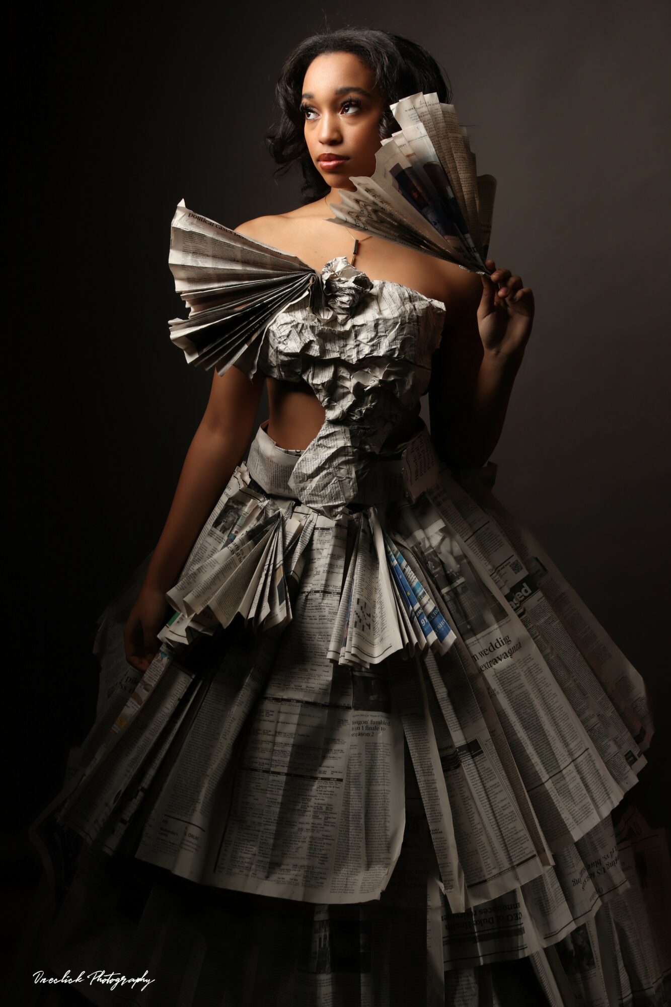 Woman in an elaborate dress made of newspaper pages, with large newspaper fans on her shoulders, against a dark background.
