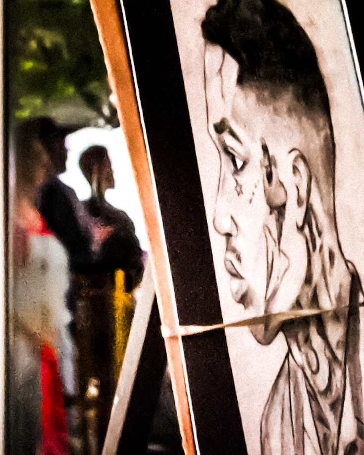 Sketch of a person's face with a hairstyle, drawn on a vertical panel, with blurred people in the background.
