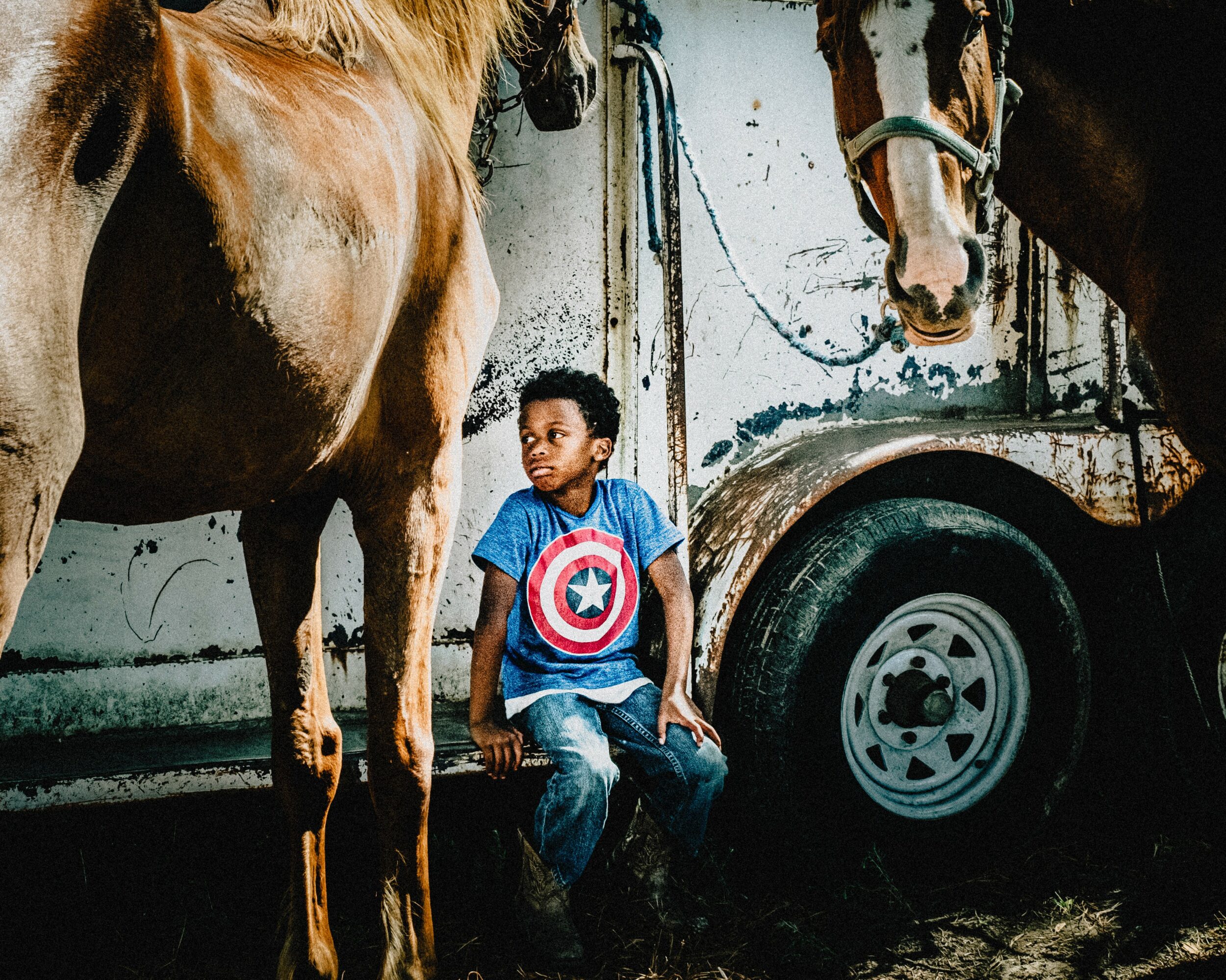 Young boy sitting between two horses near a trailer, wearing a Captain America shirt, in a rustic setting.