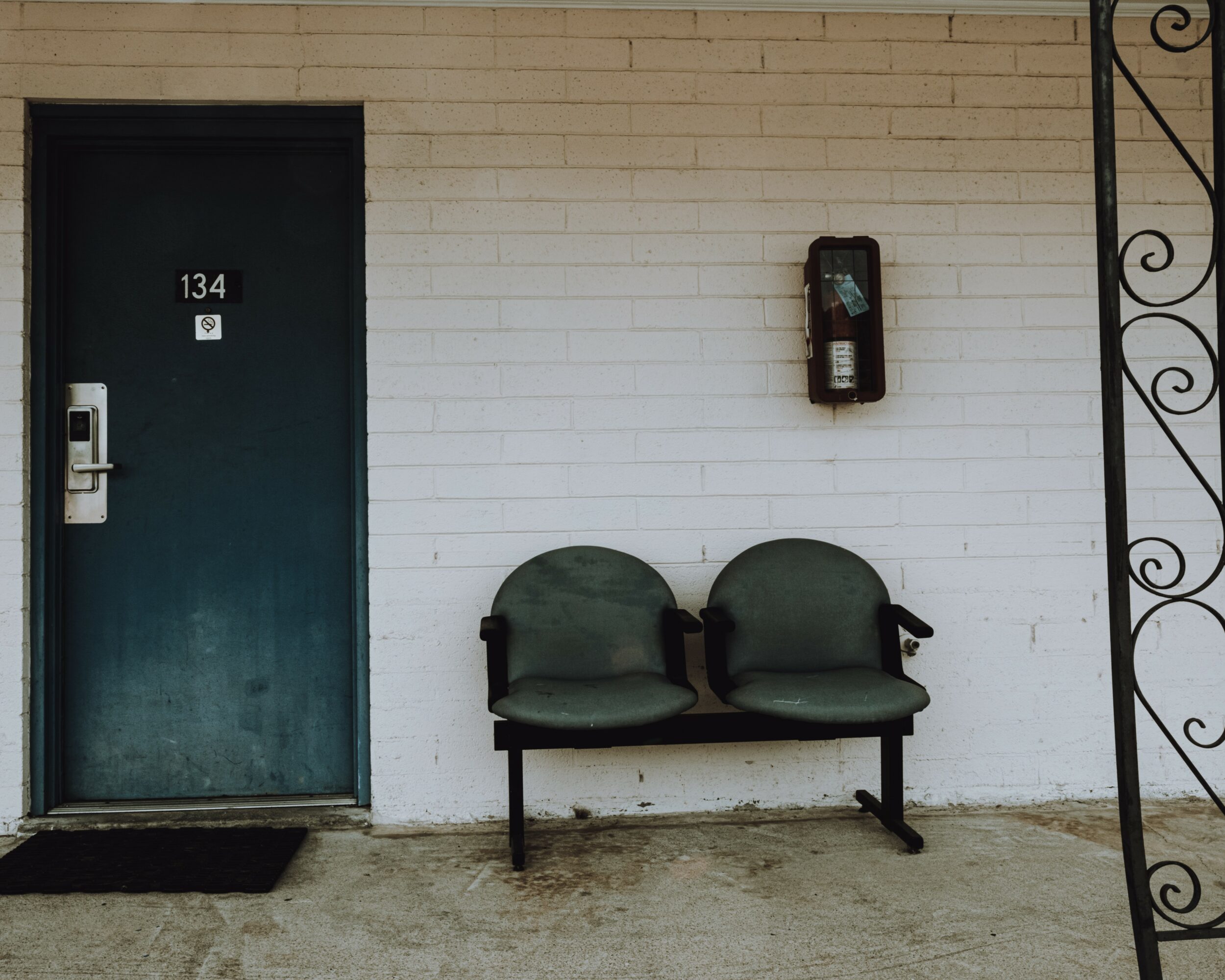 Two green chairs are placed against a white brick wall outside a building with a door labeled 134.