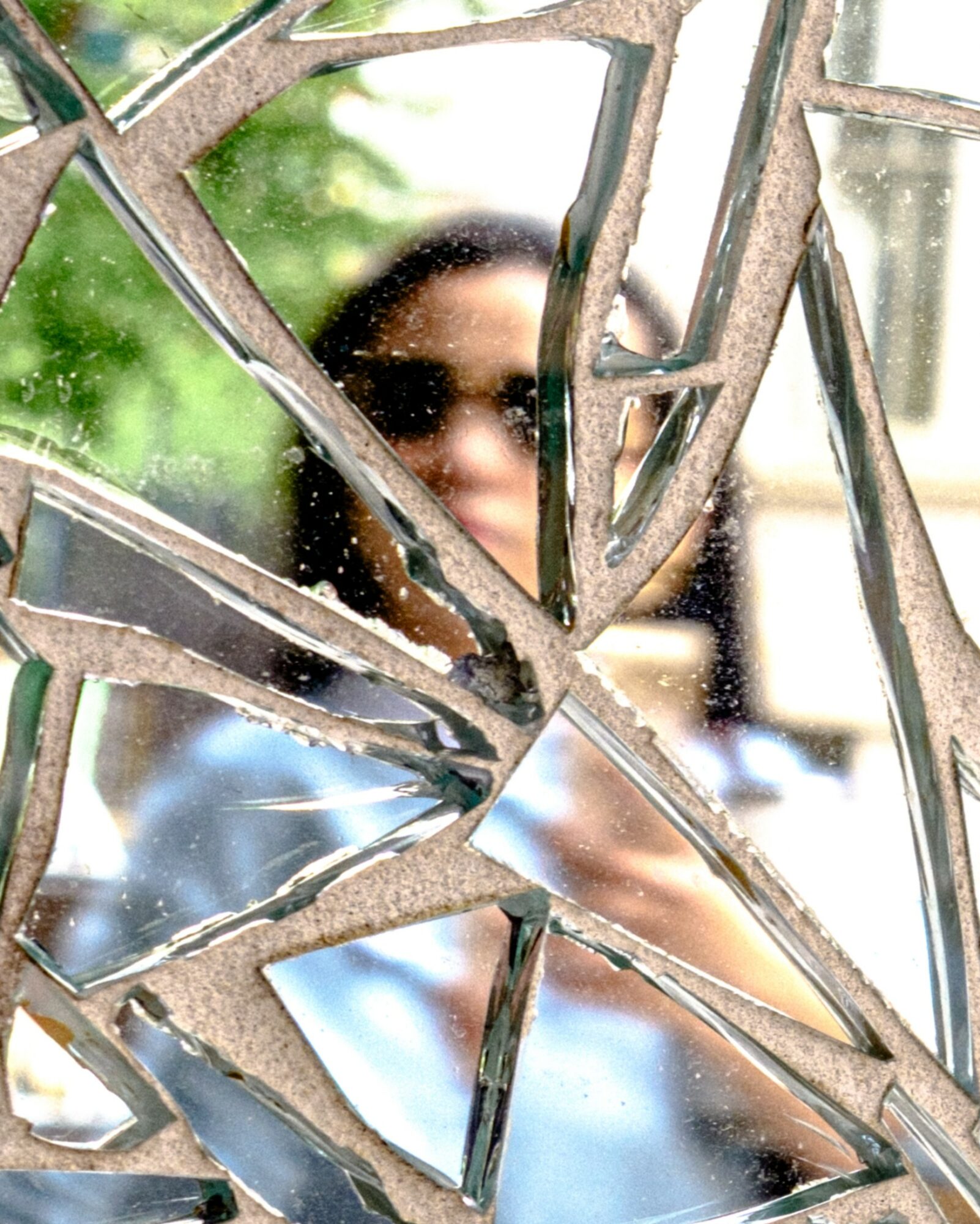 Person with dark hair and sunglasses seen through broken glass with multiple cracks and shards.