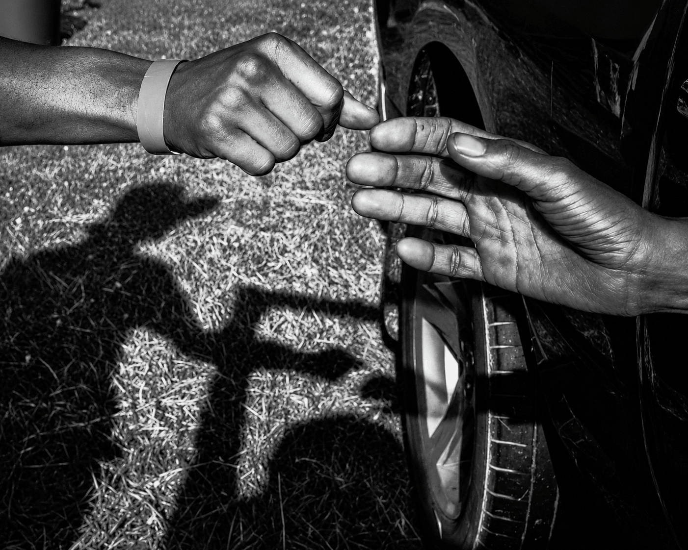 Two hands, one with a wristband, exchange a small object near a car, with shadows cast on the ground.