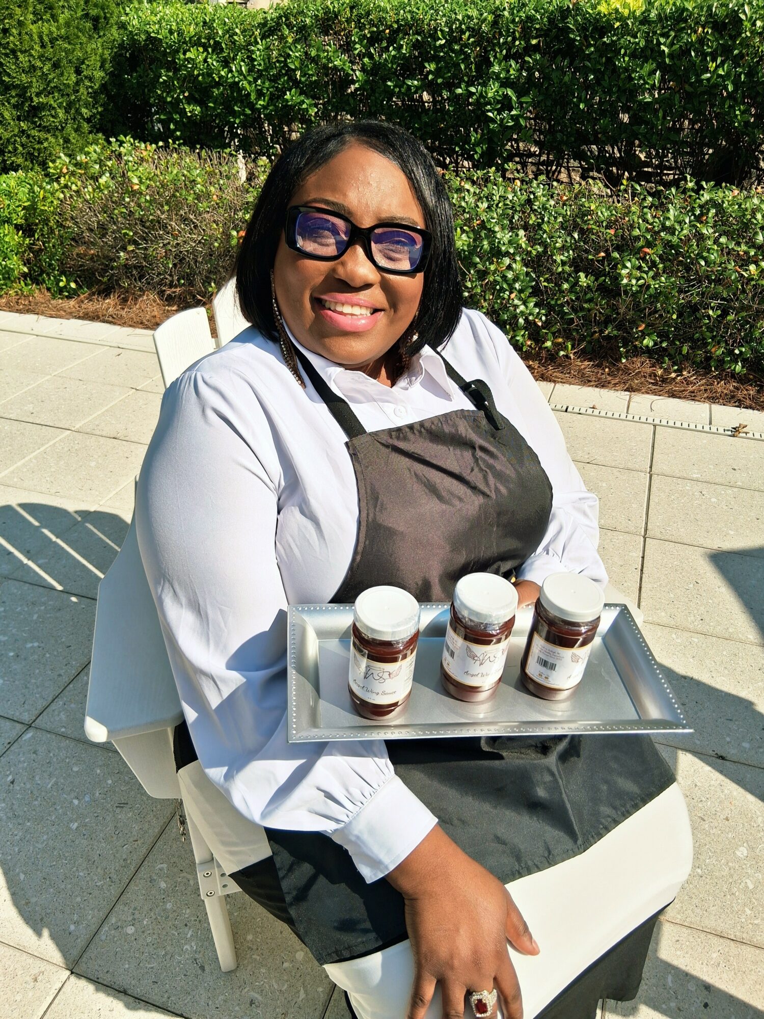 Smiling woman with glasses holding a tray of four jars outdoors on a sunny day.
