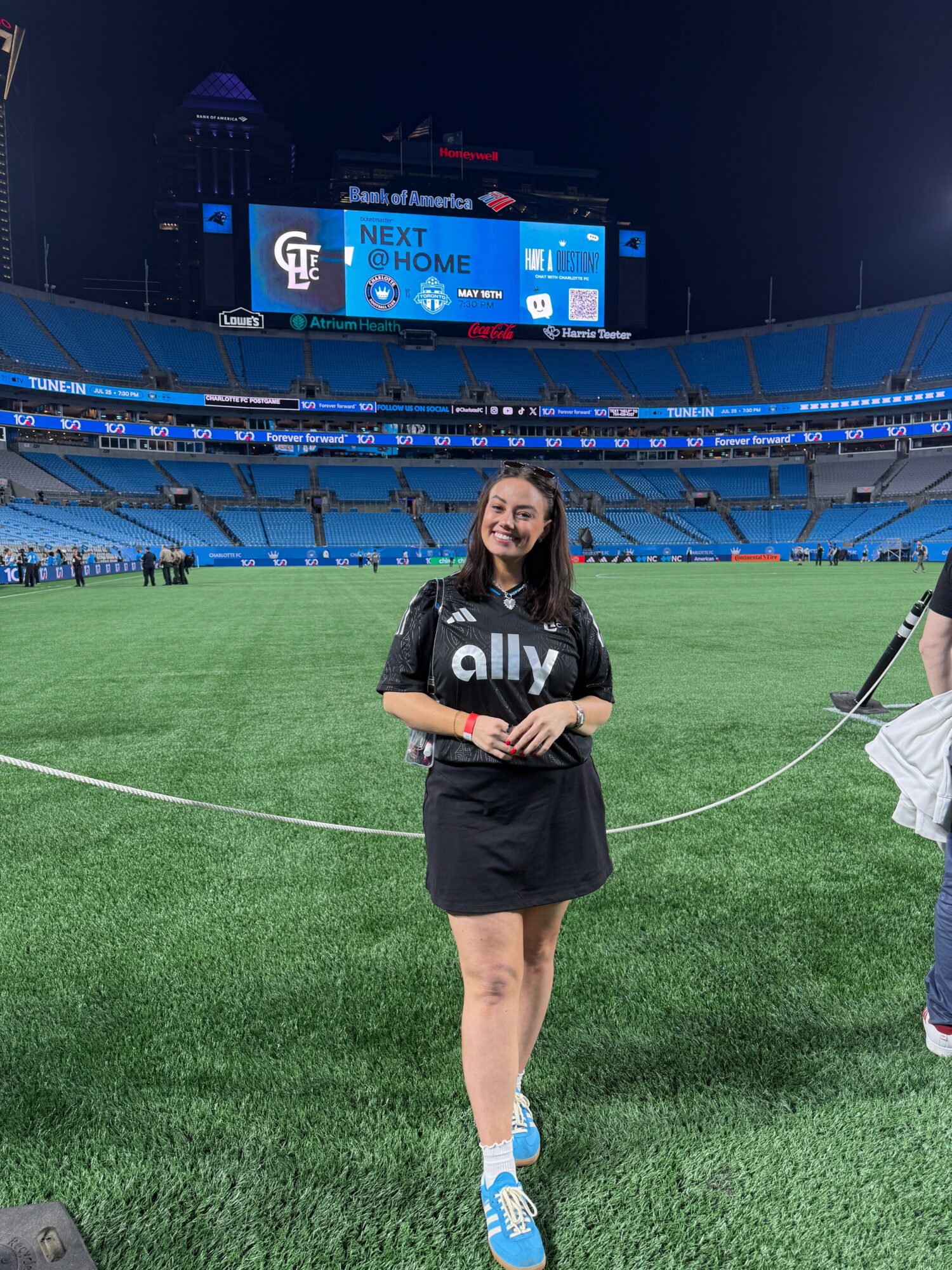Young woman standing on a baseball field at night, smiling, wearing a black shirt with 'ally' logo, blue shoes.