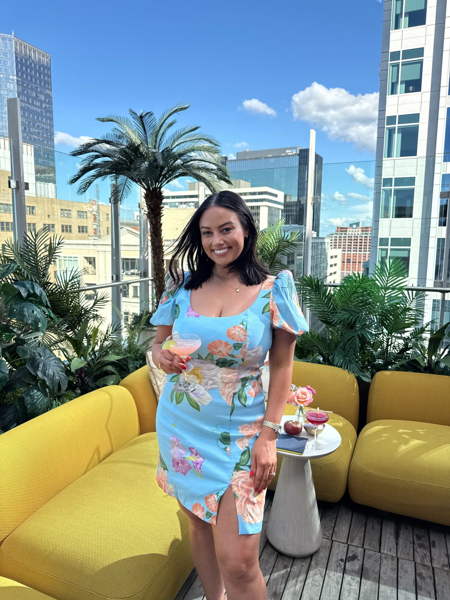 Woman in a colorful dress standing outdoors on a balcony with yellow seating and greenery, city buildings in background.