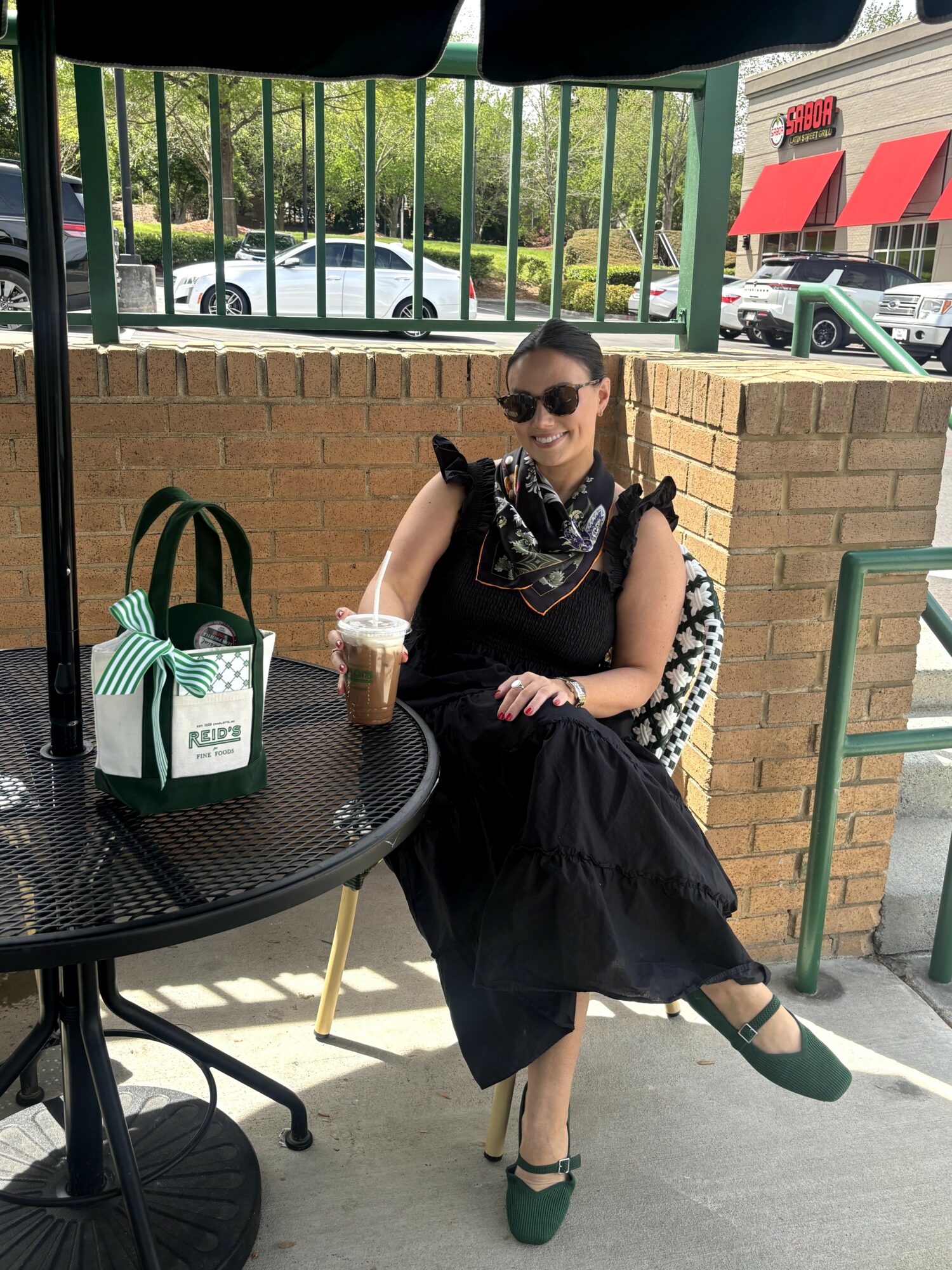 Woman sitting outdoors at a table with a drink, wearing sunglasses and a black dress, smiling.