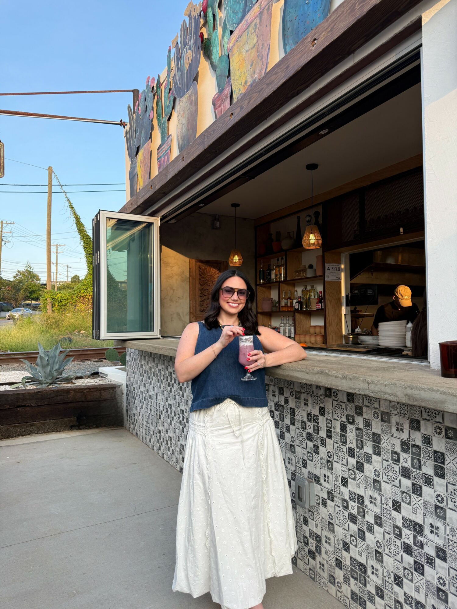 Woman with sunglasses holding a drink outside a cafe with a mosaic counter, colorful mural, and open window.