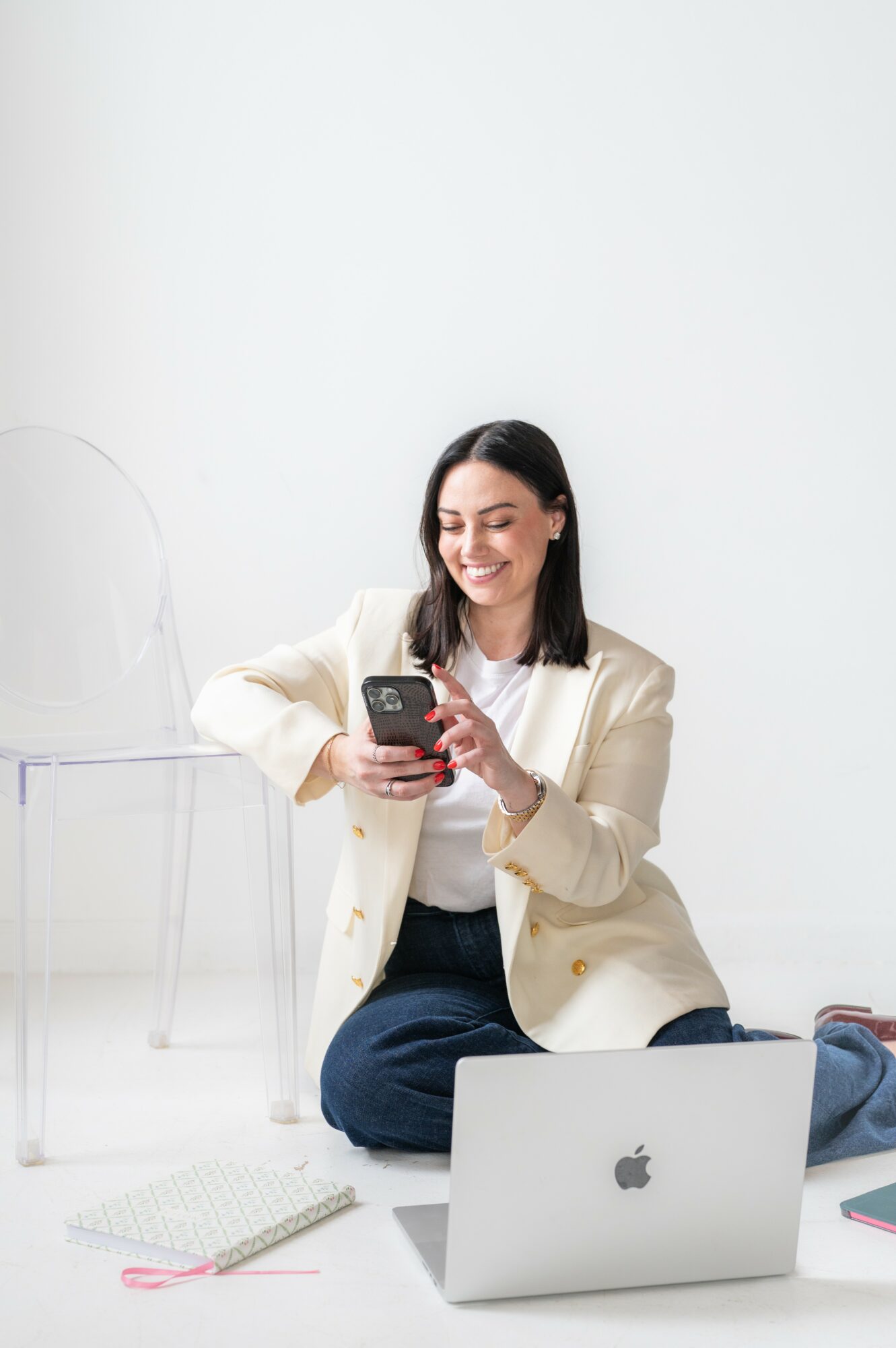 Woman sitting on the floor using a smartphone, with a laptop and notebook nearby, against a plain white background.