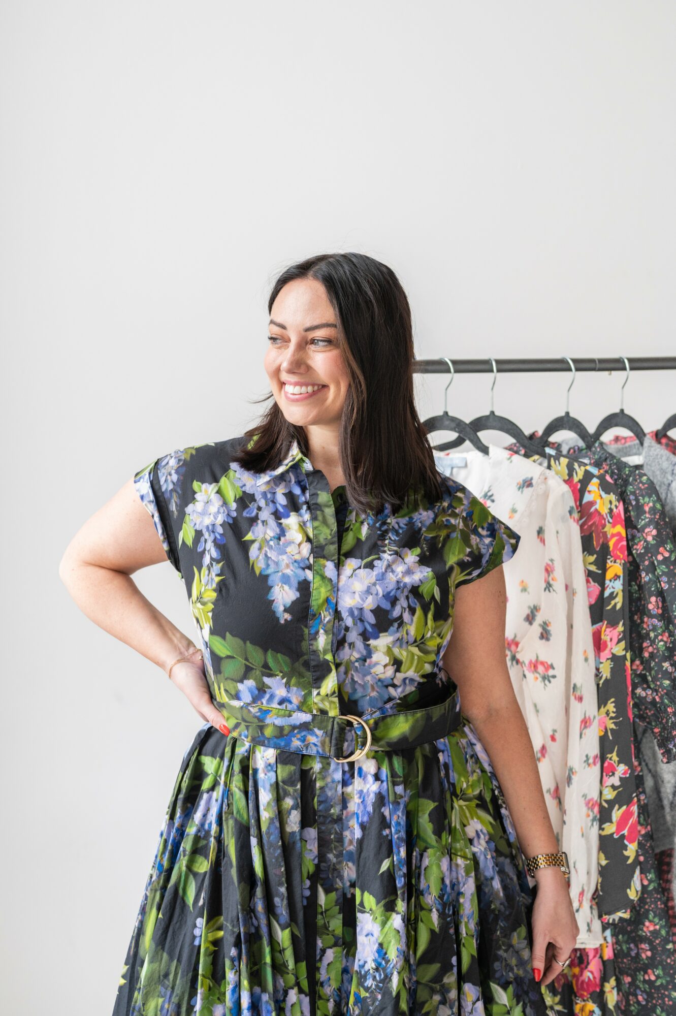 Smiling woman with dark hair wearing a floral dress, standing in front of a clothing rack with various garments.