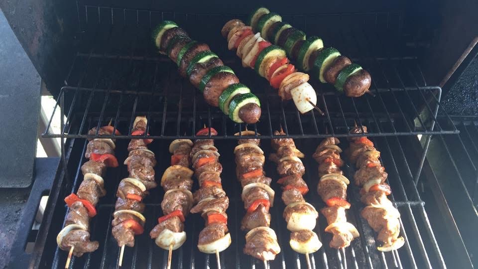 Skewered meat and vegetable kebabs cooking on a grill, with some skewers at the top and others at the bottom.