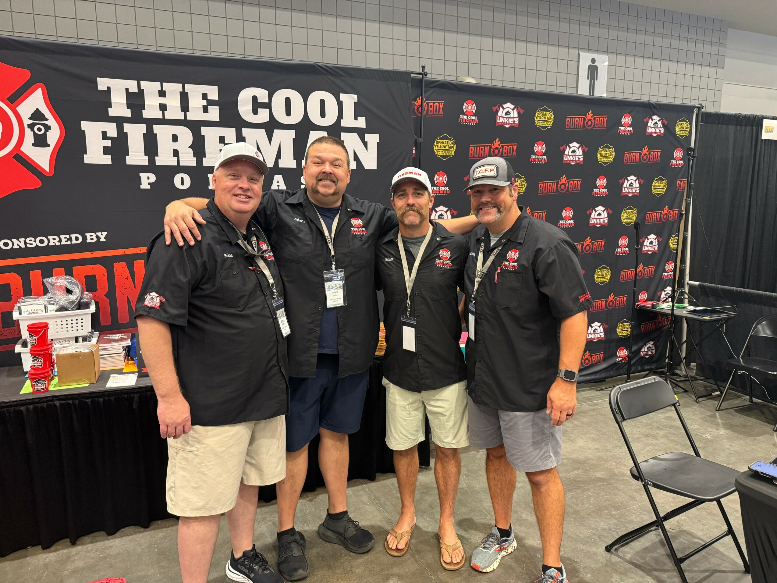 Four men in black shirts and shorts standing together at a fireman event, smiling, with a backdrop and tables behind them.