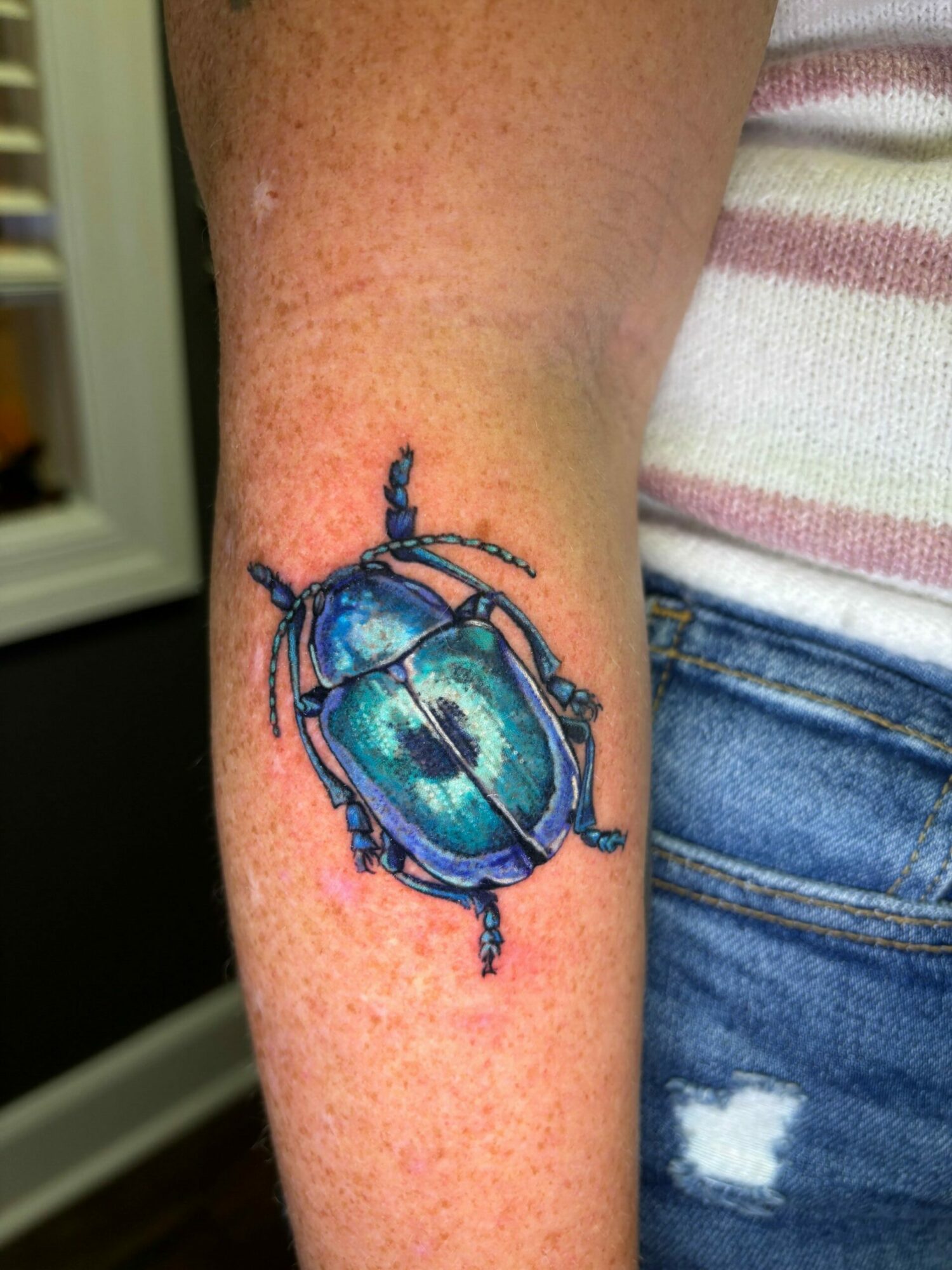 Colorful beetle tattoo on person's arm, with detailed blue, green, and black design, visible skin and clothing, 20 words.