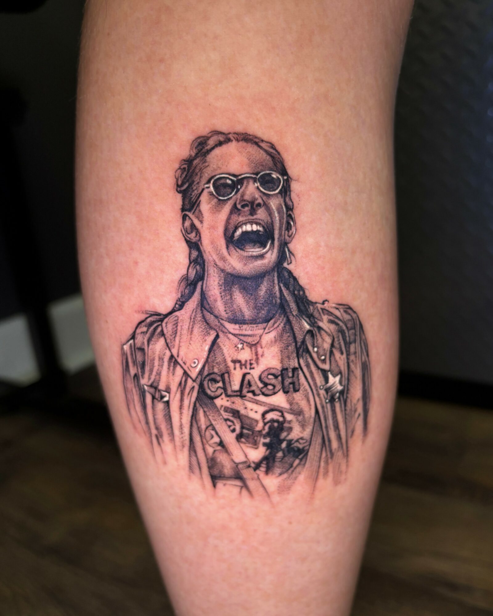 Tattoo of a person with glasses and long hair, shouting, wearing a jacket and a shirt with a band logo.