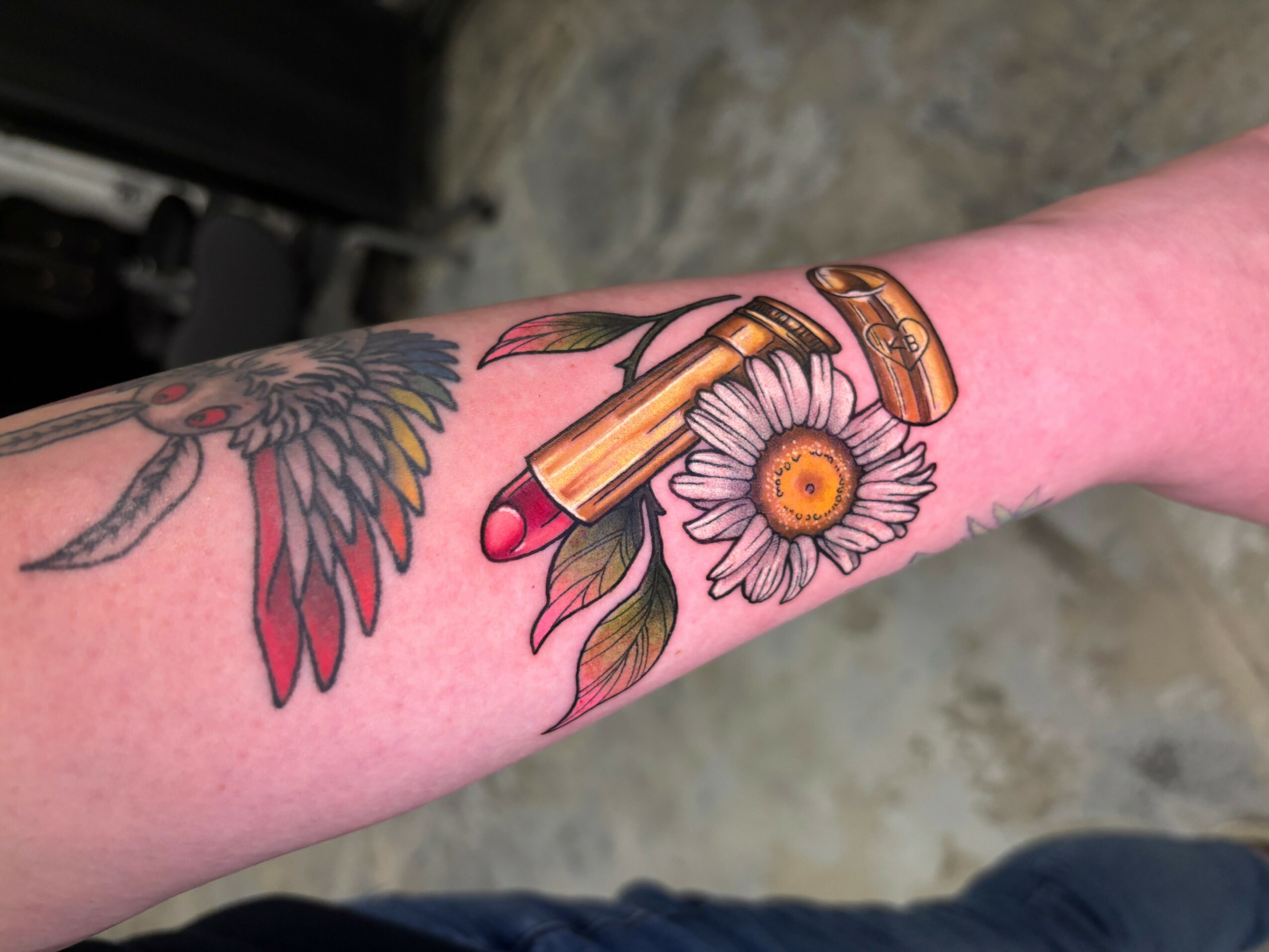Tattoo of a lipstick with a daisy flower and leaves on a person's forearm.