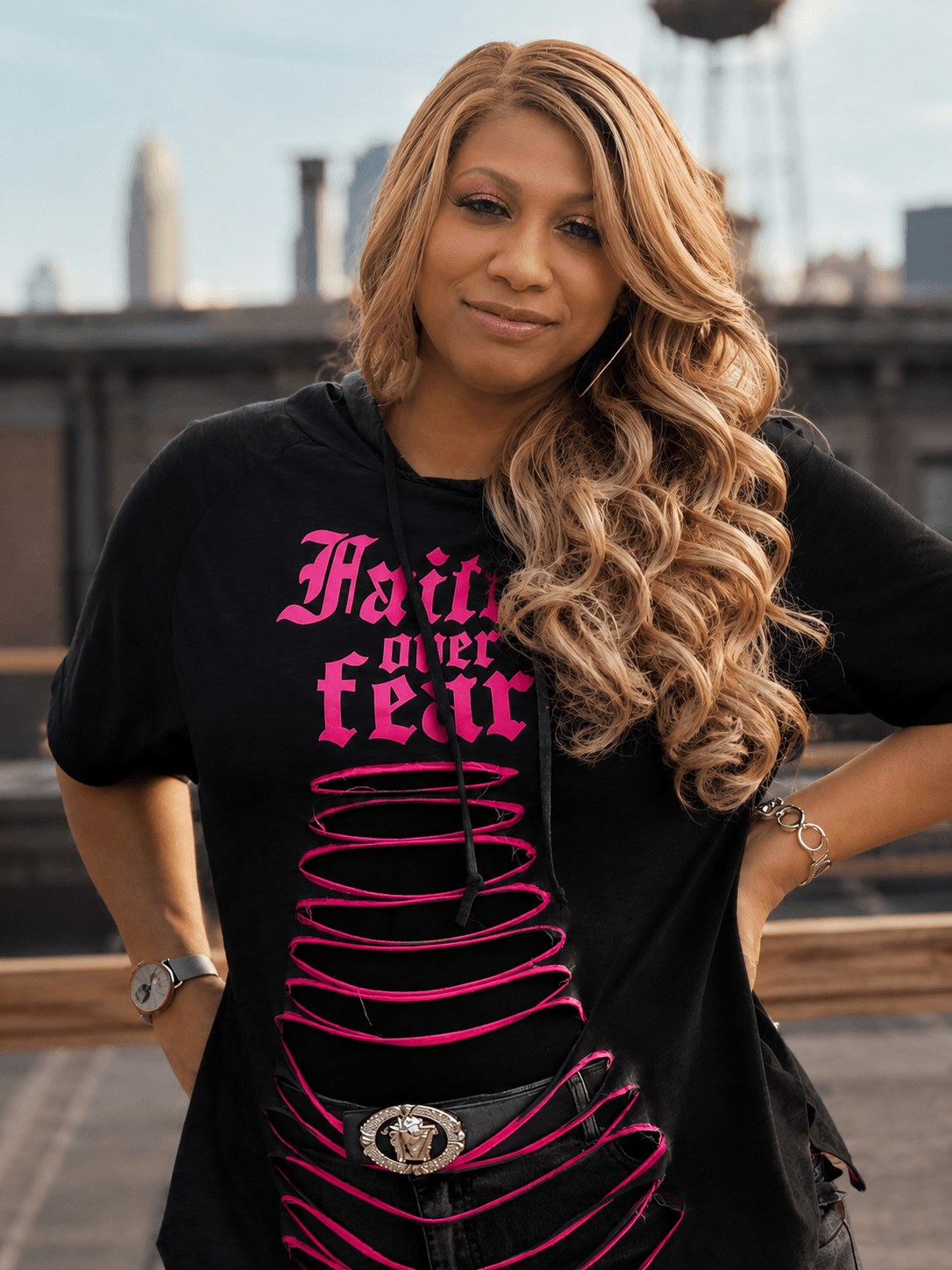 Woman with long wavy hair wearing a black T-shirt with pink text and design, standing outdoors with city buildings in background.
